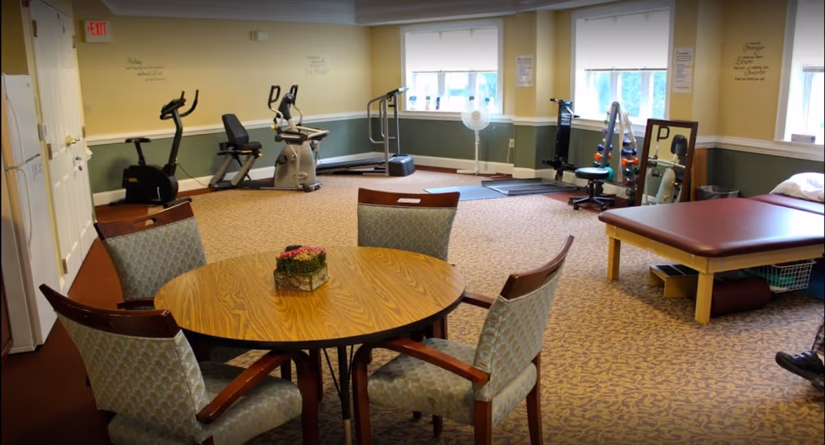 A room with exercise equipment including stationary bikes and a treadmill, a round wooden table with four upholstered chairs, a therapy or massage table, and a rack of dumbbells. The room has beige walls with green wainscoting, carpeted floor, and three windows letting in natural light.