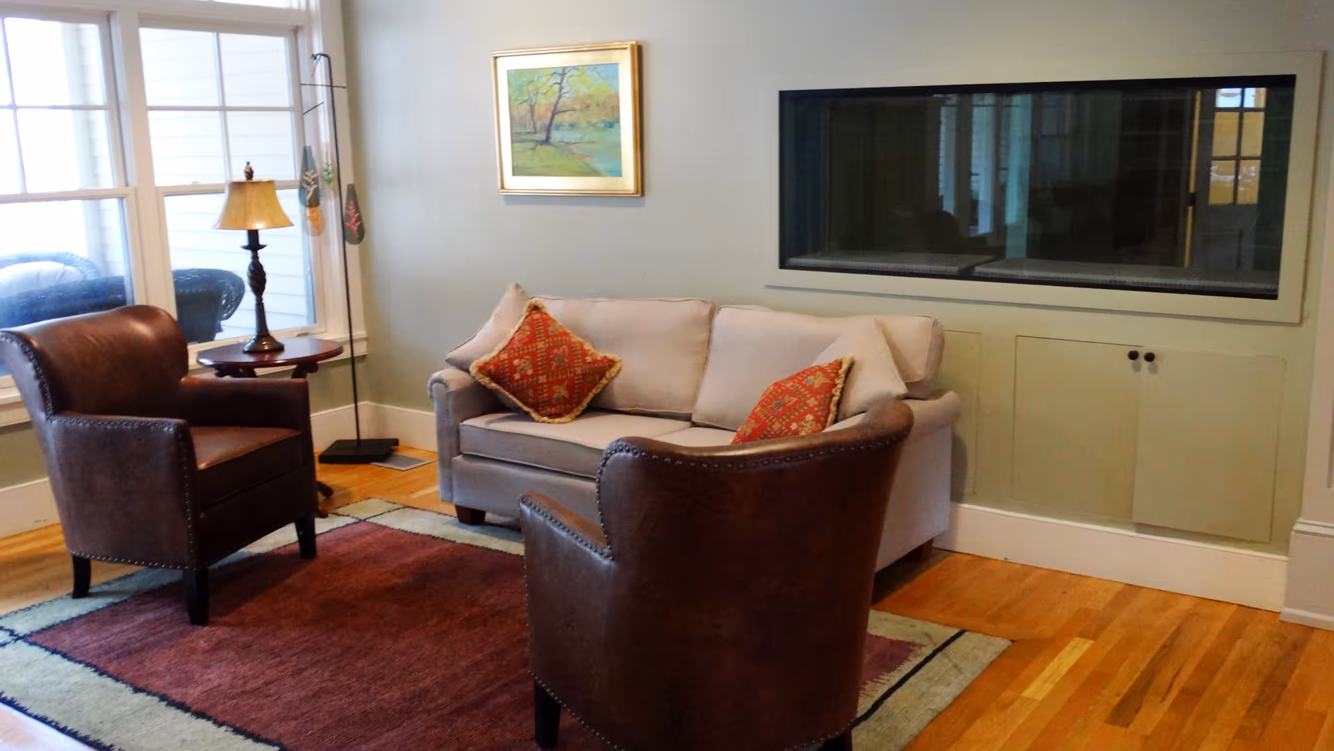 Cozy living room with a sofa, two leather armchairs, an area rug, side table with a lamp, and a framed painting on the wall.