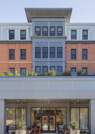 Exterior front view of a multi-story senior living facility building with a combination of brick and gray paneling. The entrance features glass doors and is flanked by potted plants and seating areas. The building has multiple windows and a modern architectural design.