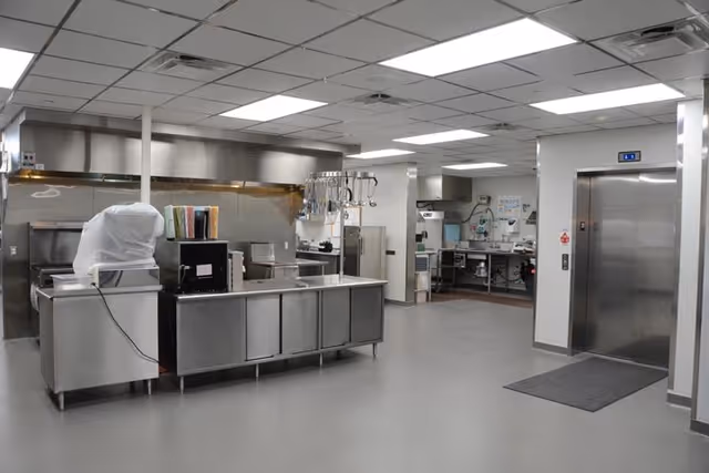 A clean, industrial kitchen area with stainless steel counters, appliances, and cooking equipment. There is an elevator on the right side of the image and various kitchen tools hanging above the counter. The ceiling has fluorescent lighting and the floor is a smooth, light gray surface.