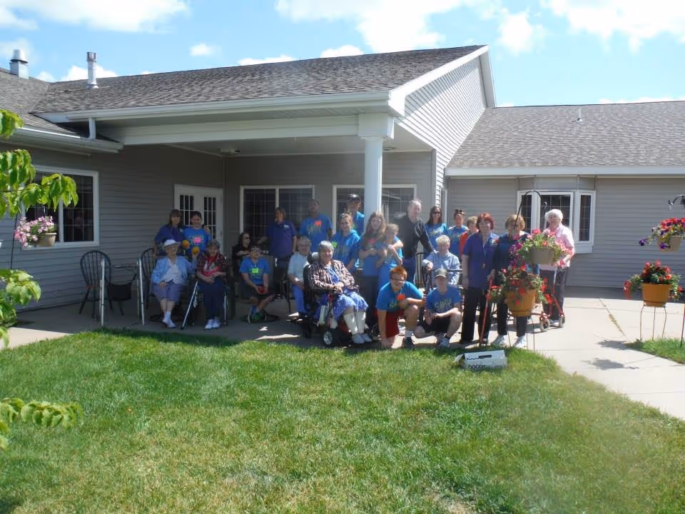 A group of elderly residents and staff members gathered outside a single-story senior living facility under a covered patio. The building has gray siding and white trim, with hanging flower baskets and green grass in the foreground. The group includes people sitting in chairs and wheelchairs, as well as standing individuals, all enjoying a sunny day.