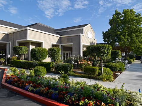 Exterior view of a senior living facility building with beige siding and a dark roof, surrounded by well-maintained landscaping including trimmed bushes, colorful flowers, and a tree, under a blue sky with scattered clouds.