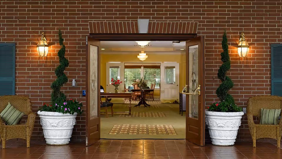 Entrance to a building with double wooden doors open, showing an interior room with a table, chairs, and a chandelier. The exterior wall is brick with two large white planters containing spiral-shaped topiary plants on either side of the doors. Two wicker chairs with green striped cushions are placed beside the planters. Two wall-mounted lantern-style lights illuminate the entrance.