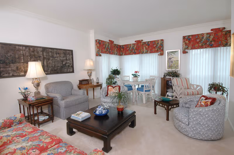 A bright living room with a variety of upholstered chairs and a floral sofa arranged around a dark wooden coffee table. The room features large windows with white vertical blinds and red floral valances. There are two table lamps on side tables, decorative plants, and framed artwork on the walls.
