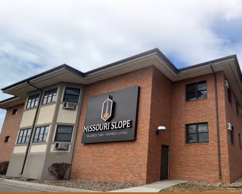 Exterior view of a two-story brick building with a large sign that reads 'MISSOURI SLOPE TAILORED CARE. INSPIRED LIVING.' The building has several windows and a partly cloudy sky above.