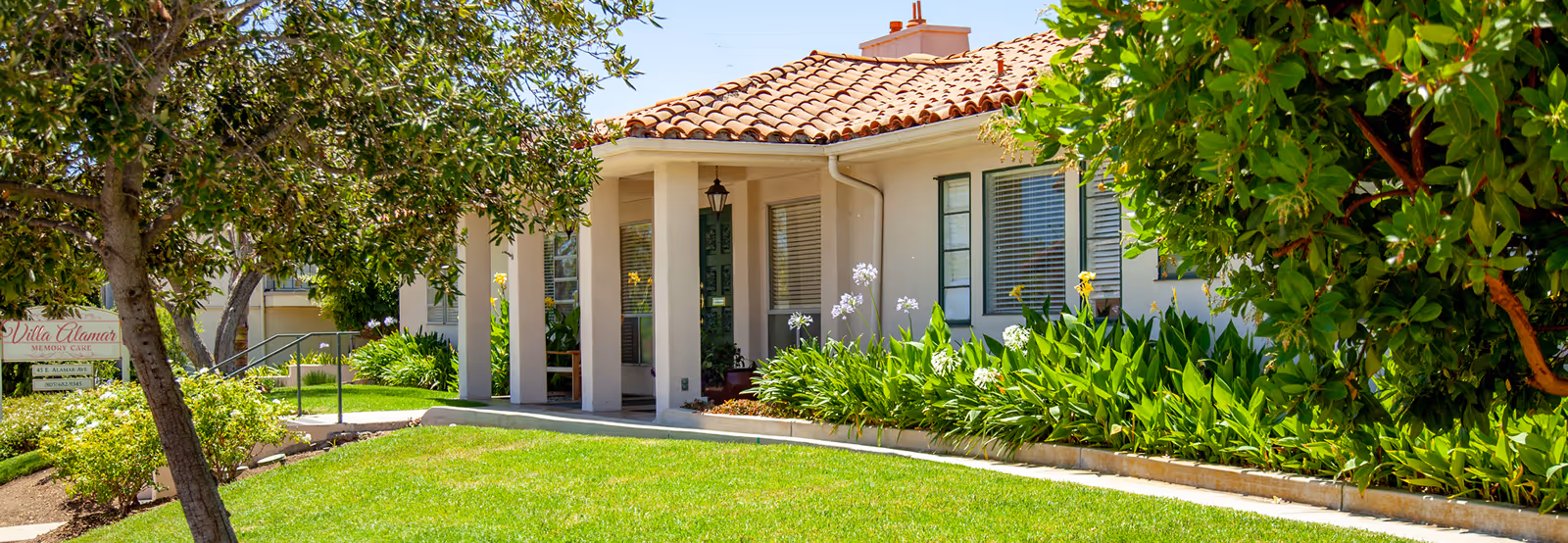 Exterior view of Villa Alamar facility showing a single-story building with a tiled roof, a green door, windows with blinds, and a well-maintained garden with green grass, bushes, and trees.