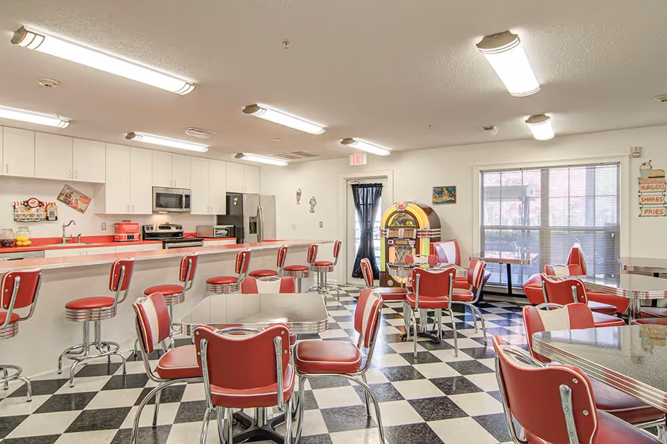 A retro-style dining area with red and white chairs and stools, checkered black and white floor tiles, a long counter with a sink and kitchen appliances, and a vintage jukebox near a window with blinds.