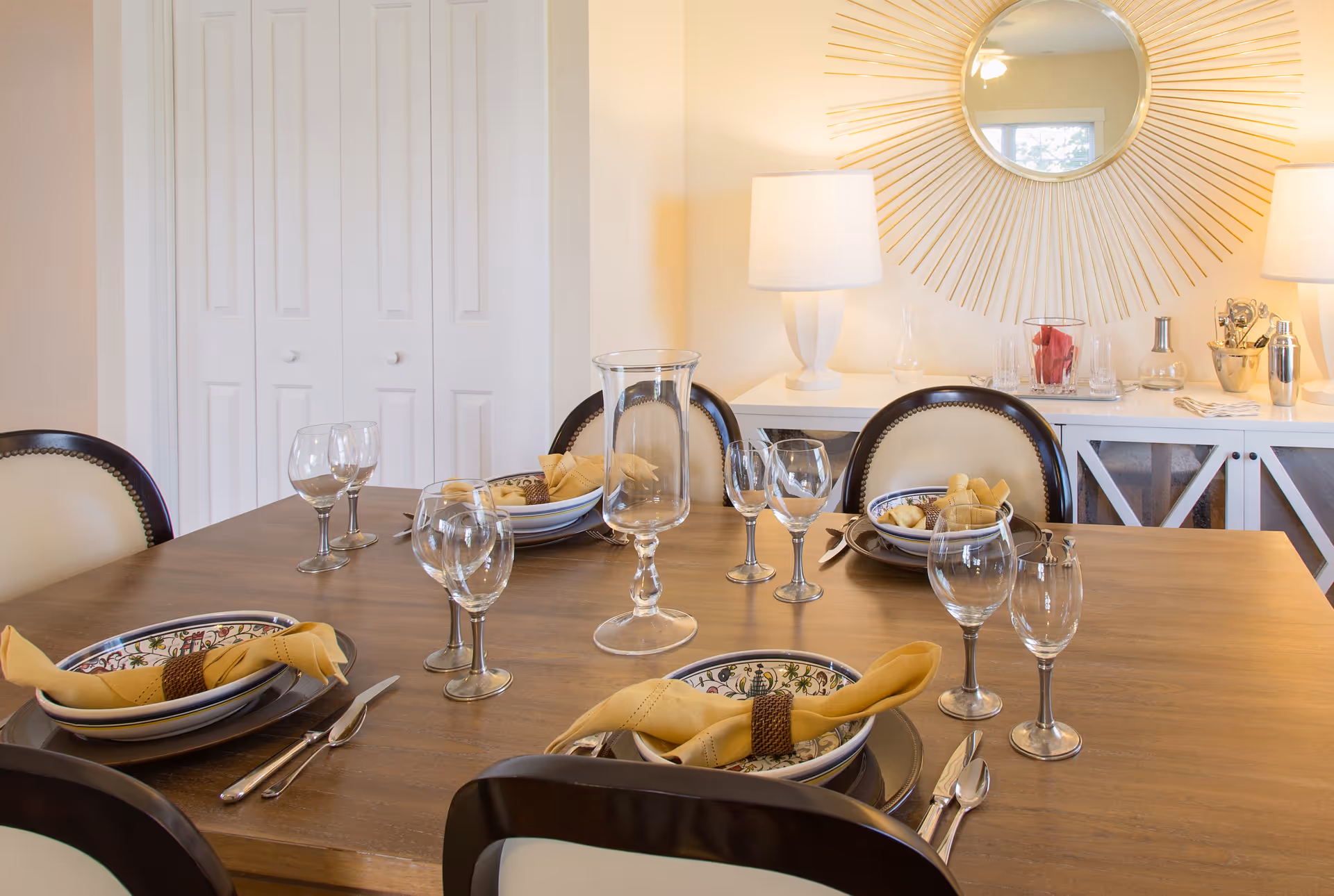 A dining room table set for four with decorative plates, yellow napkins, and multiple wine glasses. The table is wooden with dark chairs featuring light upholstery. In the background, there is a white sideboard with two lamps, a round sunburst mirror, and various glassware and decorative items.