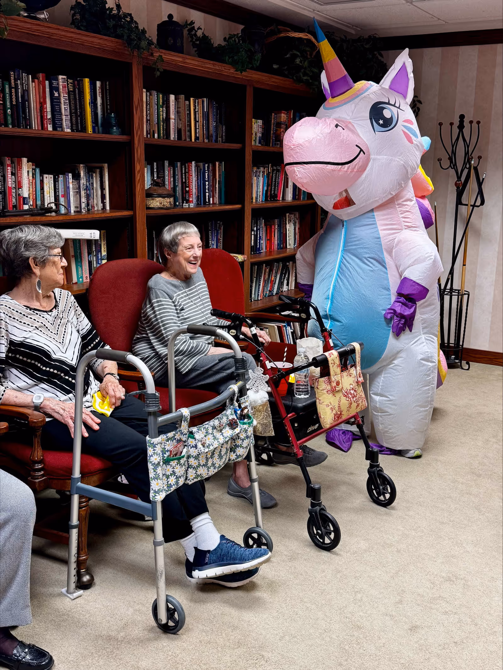 Two elderly women sitting in red chairs in a room with bookshelves filled with books behind them. Both women have walkers in front of them. A person dressed in a large, colorful inflatable unicorn costume stands nearby, interacting with the women. The room has beige carpet and light-colored walls with a coat rack in the corner.