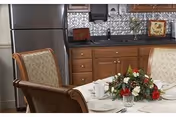 Dining area with a table set for a meal, featuring a floral centerpiece, white plates, cups, and silverware. The background shows a kitchen area with wooden cabinets, a stainless steel refrigerator, and a decorative backsplash.