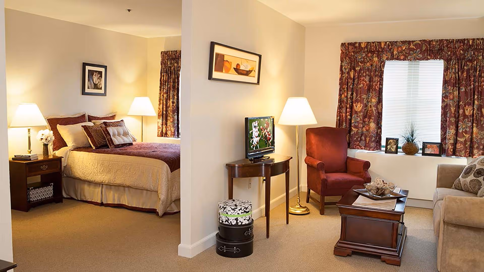 A cozy senior living facility suite with a bedroom on the left featuring a bed with multiple pillows, a nightstand with a lamp and flowers, and a living area on the right with a red armchair, beige sofa, wooden coffee table, small TV on a wooden console, floor lamp, and a window with floral curtains and decorative items on the windowsill.