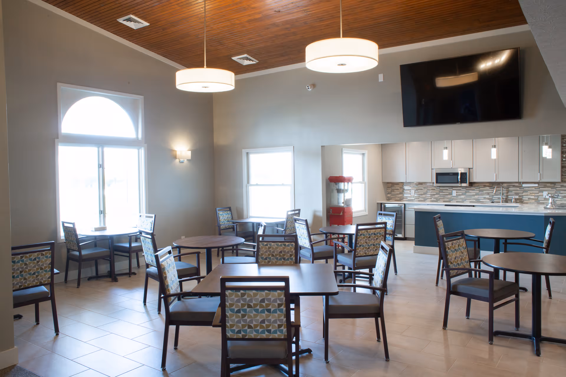 Bright communal dining area with multiple round and square tables and chairs, a kitchen counter, popcorn machine, and a wall-mounted TV.