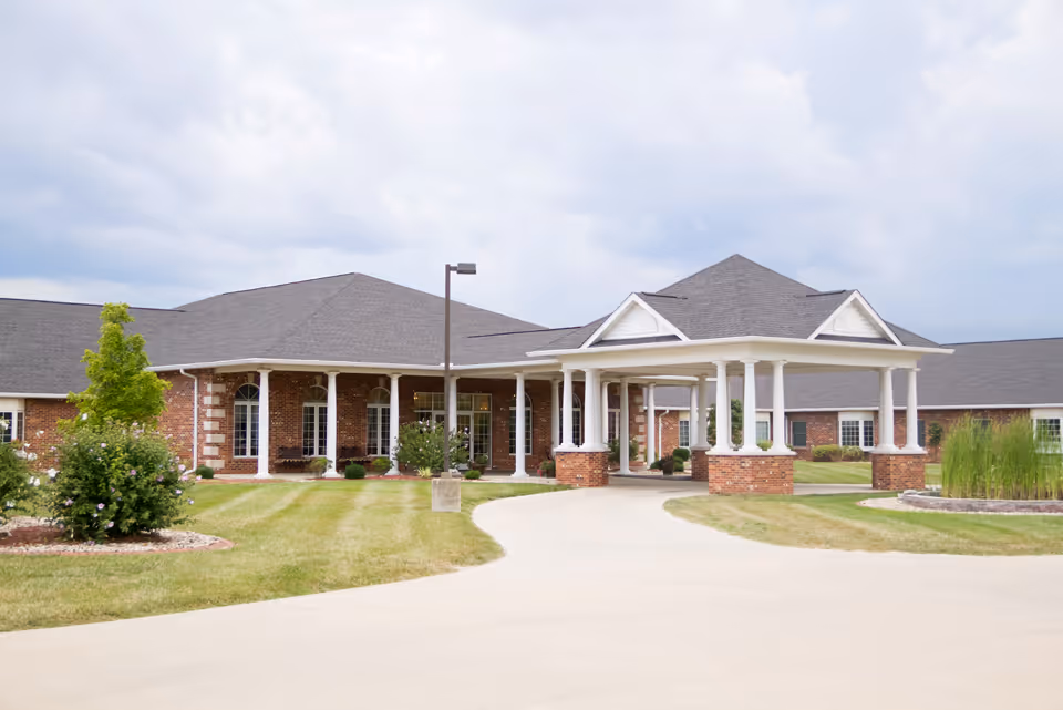 Front exterior view of a single-story brick building with a covered entrance supported by white columns, surrounded by a well-maintained lawn and landscaping under a cloudy sky.