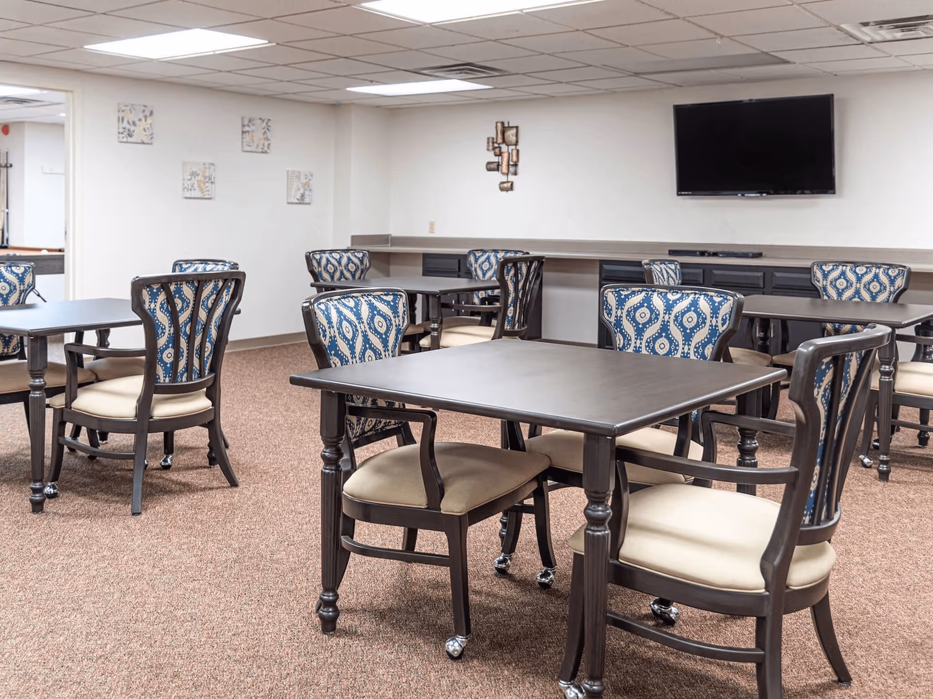 A dining area with multiple square tables and chairs featuring blue patterned backs and beige seats. The room has beige carpet, white walls with minimal decorations, a mounted flat-screen TV, and a long counter with cabinets underneath along one wall.