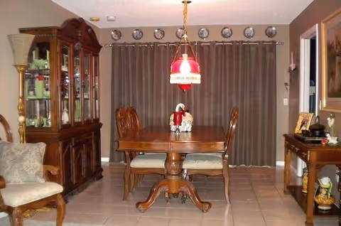 A traditional dining room with a wooden dining table and four chairs. A red and white hanging light fixture is above the table. Behind the table are brown curtains covering a window. To the left is a wooden china cabinet with glass doors displaying dishes and glassware. To the right is a wooden sideboard with decorative items and a mirror above it. The floor is tiled in a light color.