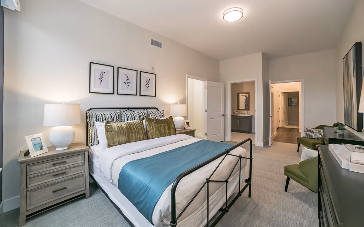 Spacious, modern bedroom with a metal-framed bed dressed in teal and white linens, bedside tables with lamps, framed wall art, and a seating area.