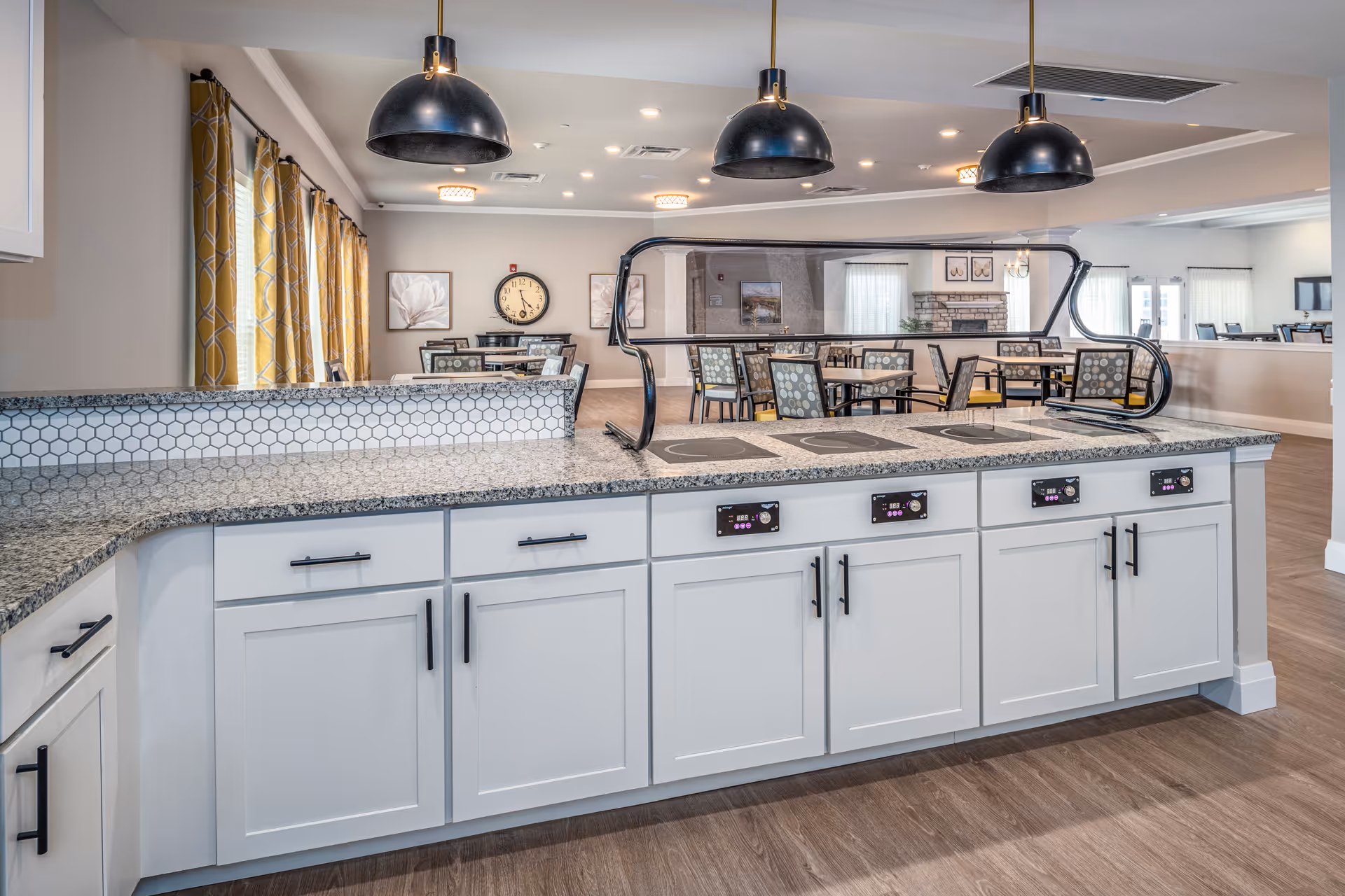 A modern kitchen area with a granite countertop and white cabinets featuring black handles. There are four electric cooktops built into the counter, each with digital controls. Above the counter hang three black pendant lights. In the background, there is a dining area with tables and chairs, yellow patterned curtains on the windows, a wall clock, and framed artwork. The flooring is wood, and the space is well-lit with recessed ceiling lights.