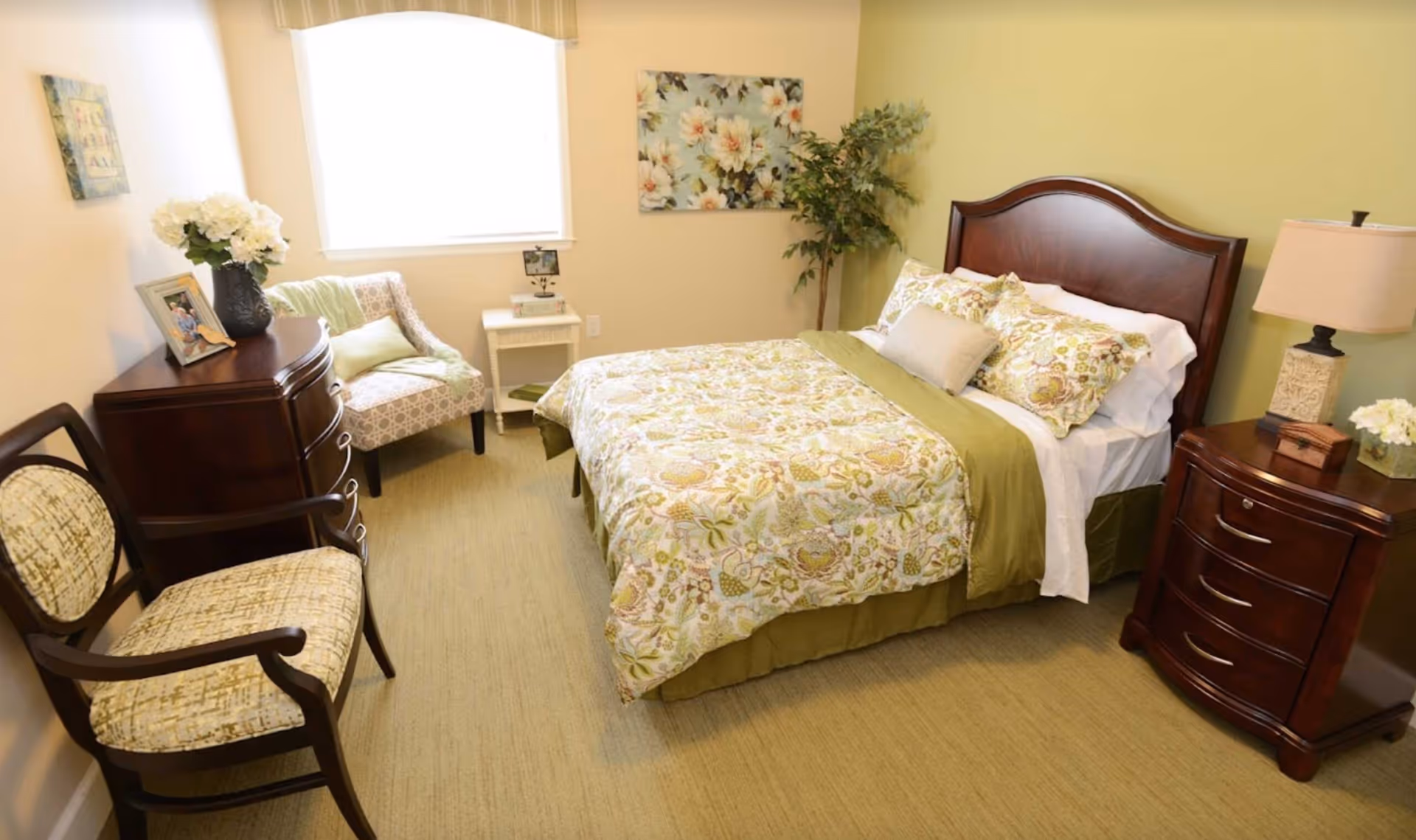 A cozy bedroom with a double bed featuring floral bedding and multiple pillows. The room has light yellow walls, a window with a valance, a wooden nightstand with a lamp and flowers, a wooden dresser with a photo frame and flowers, two upholstered chairs, and a decorative plant in the corner.