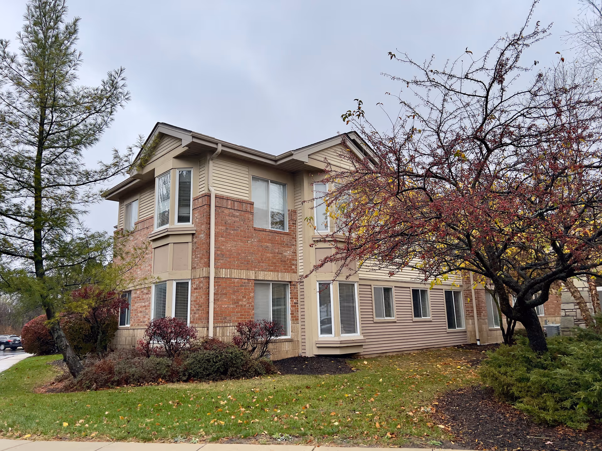 Exterior view of a two-story building with beige siding and red brick accents, surrounded by green grass, bushes, and trees with autumn leaves under a cloudy sky.