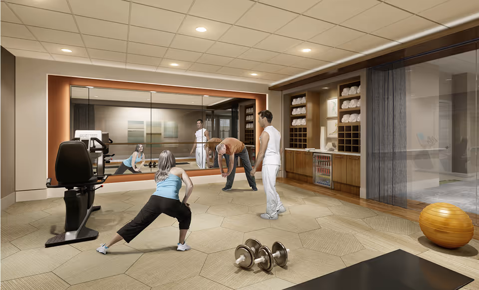 A fitness room in a senior living facility with three people stretching and exercising in front of a large mirror. The room has exercise equipment including a recumbent bike, dumbbells on the floor, and an orange exercise ball. There are shelves with neatly stacked towels and a small refrigerator in the background.