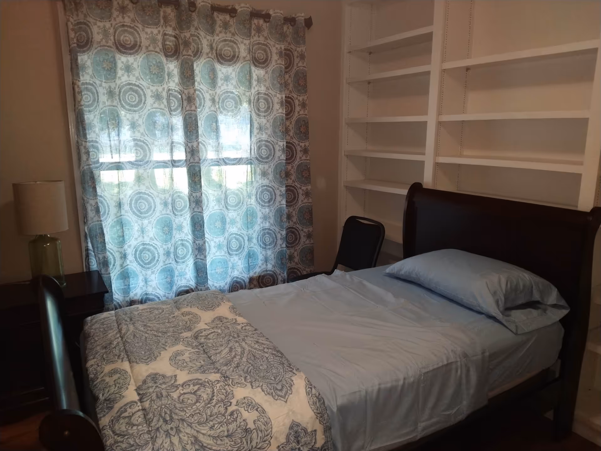 Small bedroom with a single dark-wood sleigh bed made with blue sheets, patterned curtains over a window, built-in white shelves, and a nightstand with a lamp.