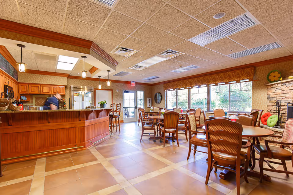 Bright communal dining and seating area with wooden tables and chairs, a serving bar, large windows, and a stone fireplace.
