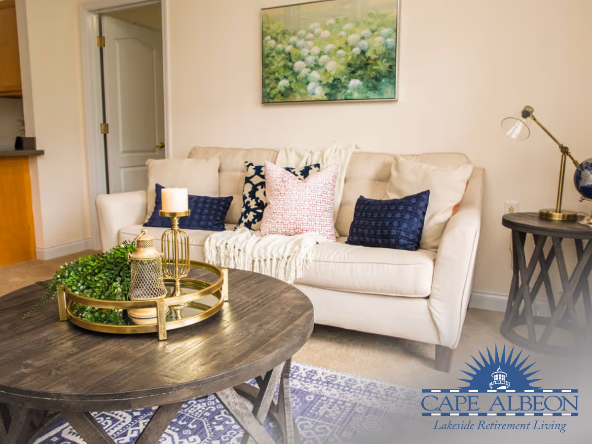 A cozy living room with a beige sofa adorned with multiple decorative pillows and a white throw blanket. In front of the sofa is a round wooden coffee table with a gold tray holding a candle, a lantern, and a green plant. A side table with a lamp and a globe is positioned to the right of the sofa. A painting of white flowers hangs on the wall above the sofa.