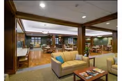 A spacious and well-lit common area in a senior living facility featuring comfortable seating with beige sofas and a wooden coffee table in the foreground. The room has wooden columns and trim, green walls, and multiple dining tables and chairs in the background. The ceiling has recessed lighting and a chandelier over the dining area.