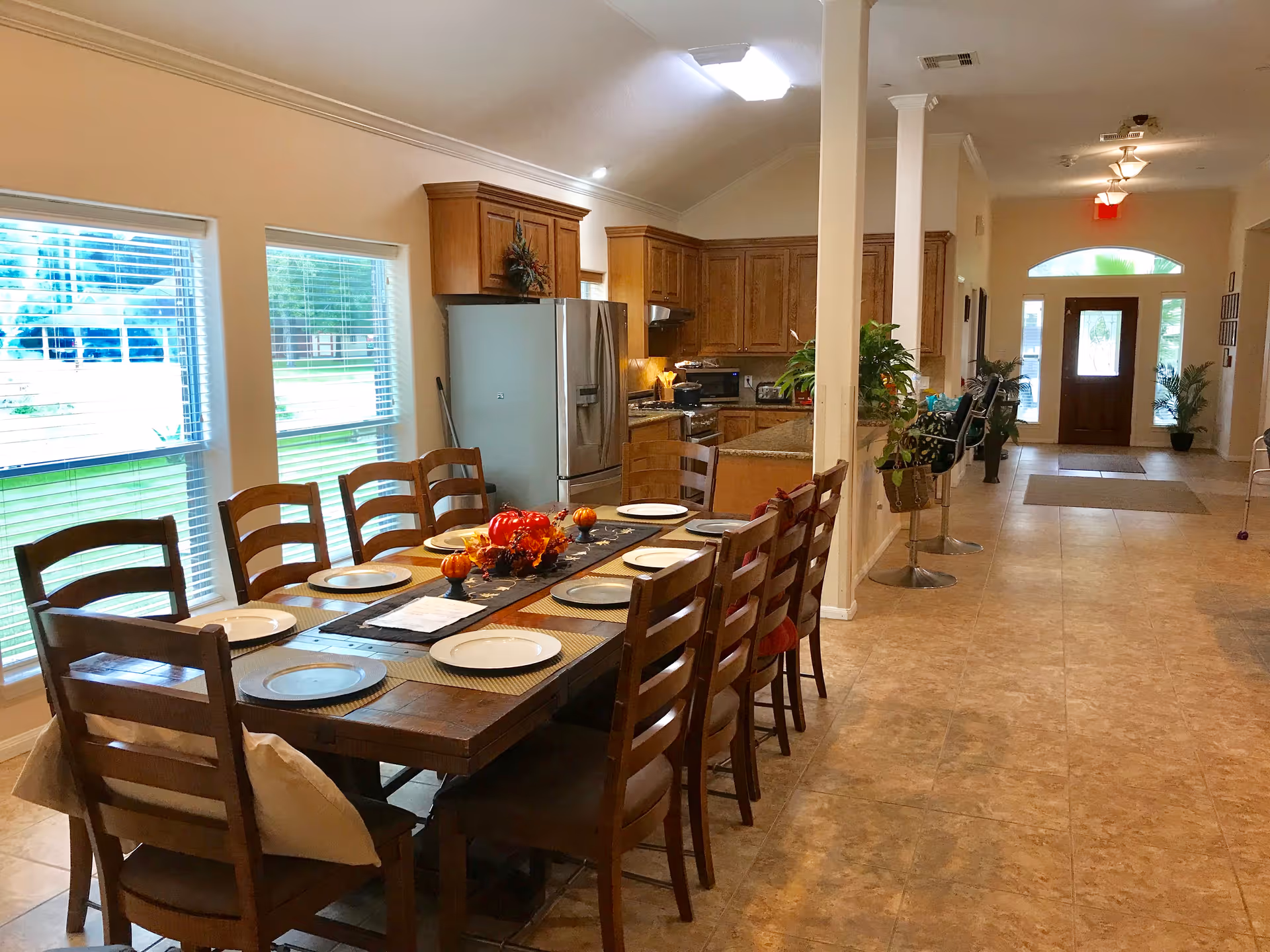 A spacious dining area with a long wooden table set with plates and autumn-themed decorations. The room features large windows with blinds, a kitchen area with wooden cabinets and stainless steel appliances, and an open hallway leading to a wooden front door with glass panels. There are plants and bar stools along the hallway.