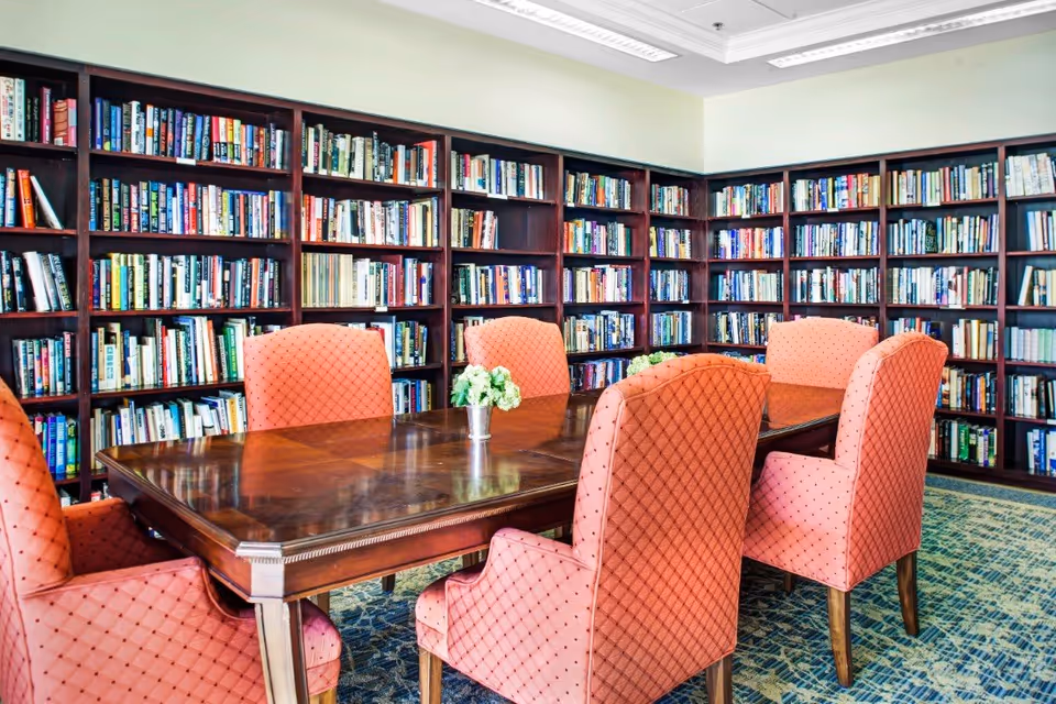 A well-lit library room with dark wooden bookshelves filled with books lining the walls. In the center, there is a polished wooden table surrounded by six upholstered chairs in a coral fabric with a diamond pattern. Small floral arrangements are placed on the table.