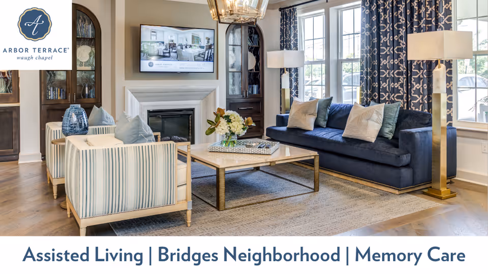 A bright and elegant living room featuring a navy blue sofa with multiple cushions, two striped armchairs, a square coffee table with a flower arrangement, two tall floor lamps, large windows with patterned curtains, and a wall-mounted TV above a fireplace. Built-in dark wood cabinets flank the fireplace.