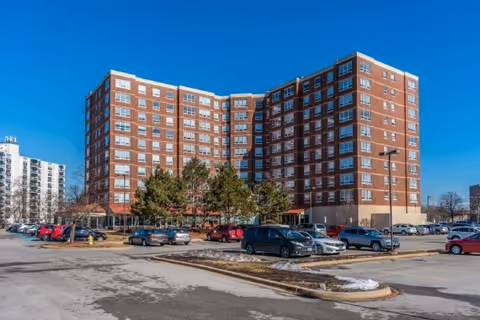 Exterior view of a large multi-story brick building with many windows, surrounded by a parking lot with several cars parked. There are some trees and patches of snow on the ground under a clear blue sky.