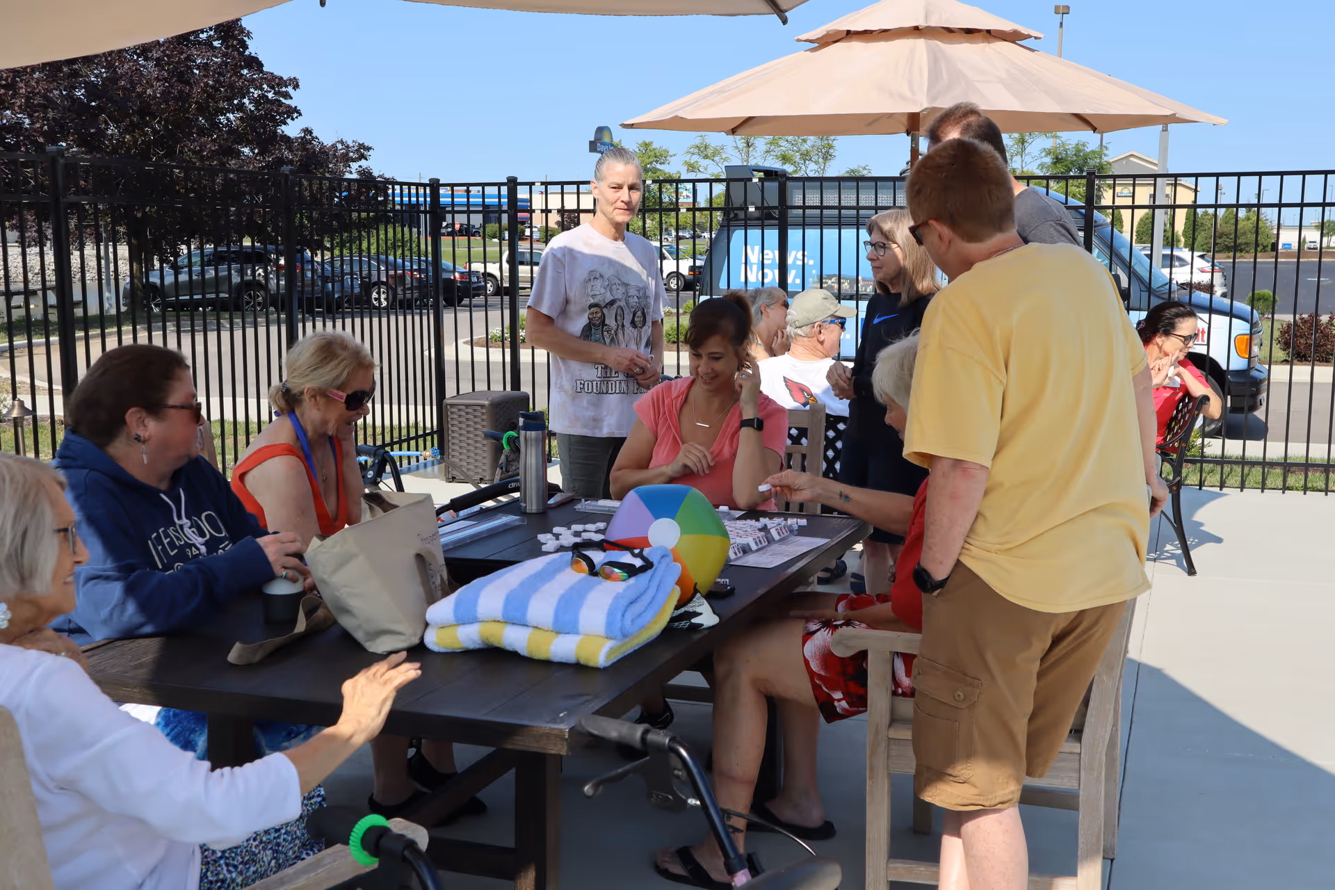 A group of senior adults and a few younger people gathered around a table outdoors under large umbrellas. They appear to be engaged in a social activity with game pieces on the table. The setting is a fenced patio area with a parking lot and vehicles visible in the background.