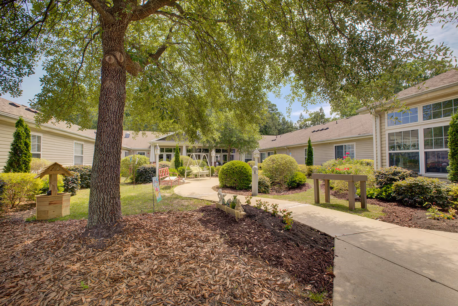 Outdoor courtyard area at Falls River Village Assisted Living featuring a large tree with green leaves providing shade, a curved concrete walkway, well-maintained bushes and flower beds, a wooden wishing well decoration, and a swing bench near the building entrance.