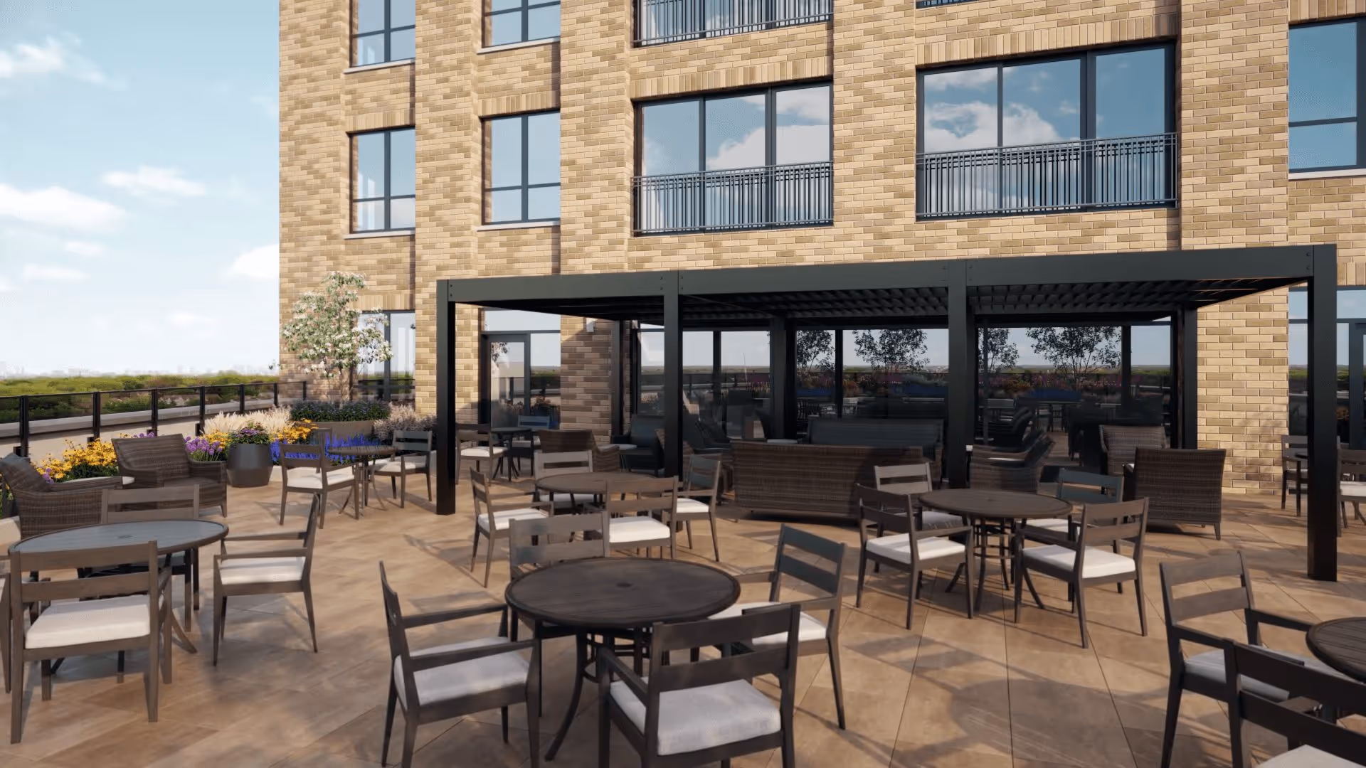 Outdoor patio area at Clarendale Six Corners with multiple round tables and chairs arranged on a tiled floor. There is a black pergola structure providing shade over some seating, and the building's brick exterior with large windows is visible in the background. Flower beds with colorful plants line the edge of the patio.