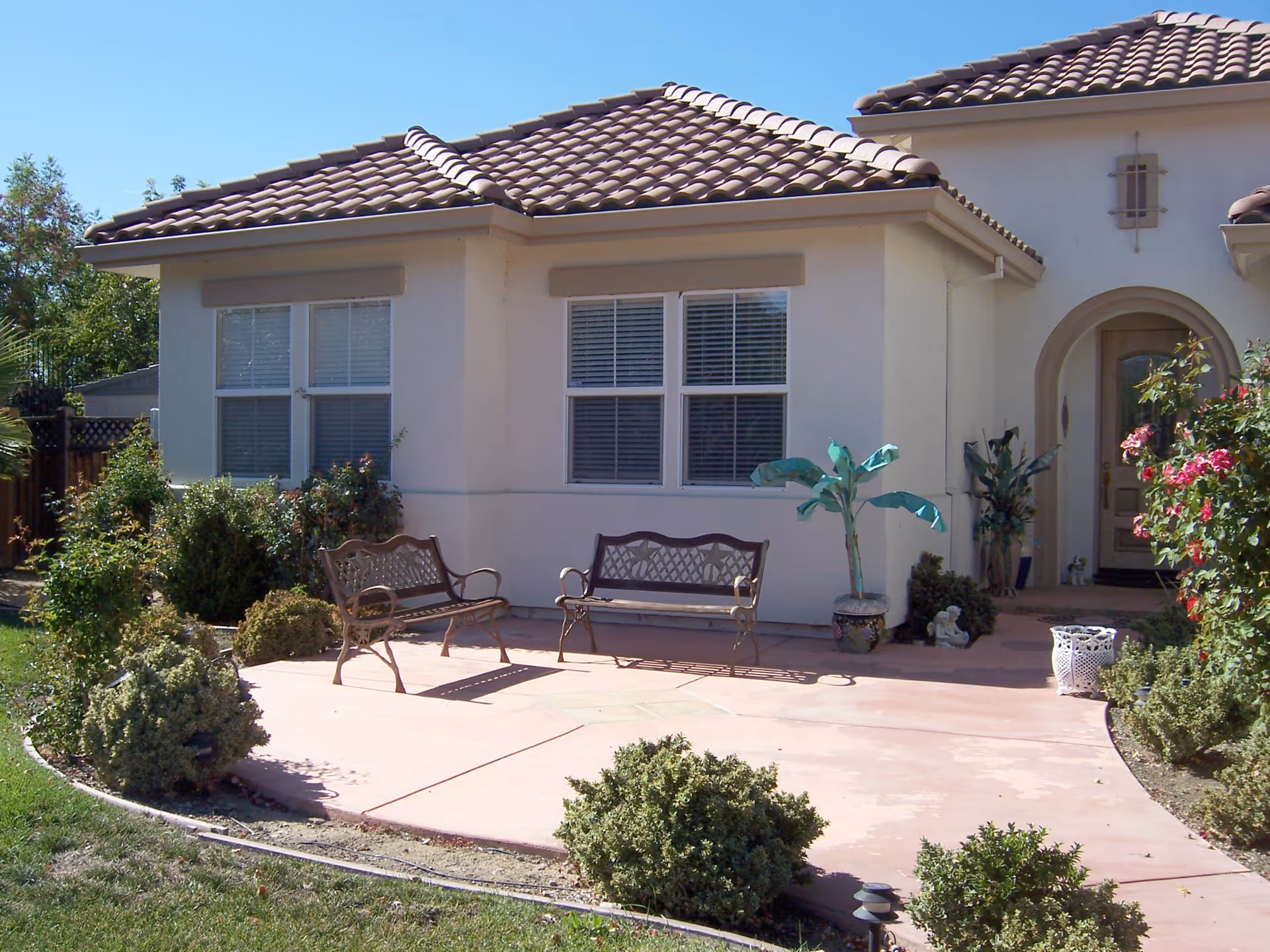 Front exterior of a stucco building with a tiled roof, a small paved patio with two metal benches and potted plants.