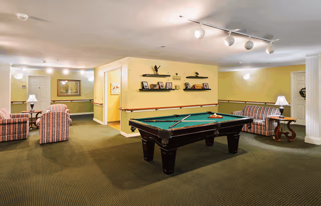 Spacious senior living common room with a pool table in the center, striped sofas and chairs, lamps, and decorative wall shelves.