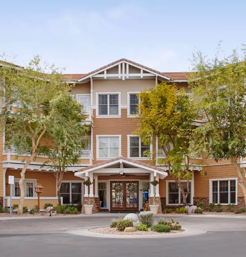 Front entrance of a three-story senior living building with a covered porte-cochere, landscaping and trees.