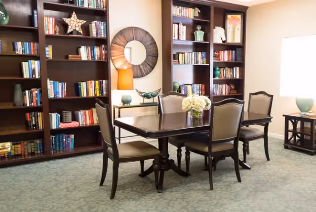 A cozy room with a dark wooden dining table surrounded by six upholstered chairs. Behind the table are built-in dark wooden bookshelves filled with books and decorative items. A round mirror and a table lamp are placed on a console table between the bookshelves. The room has a light carpet and a window with natural light coming in.