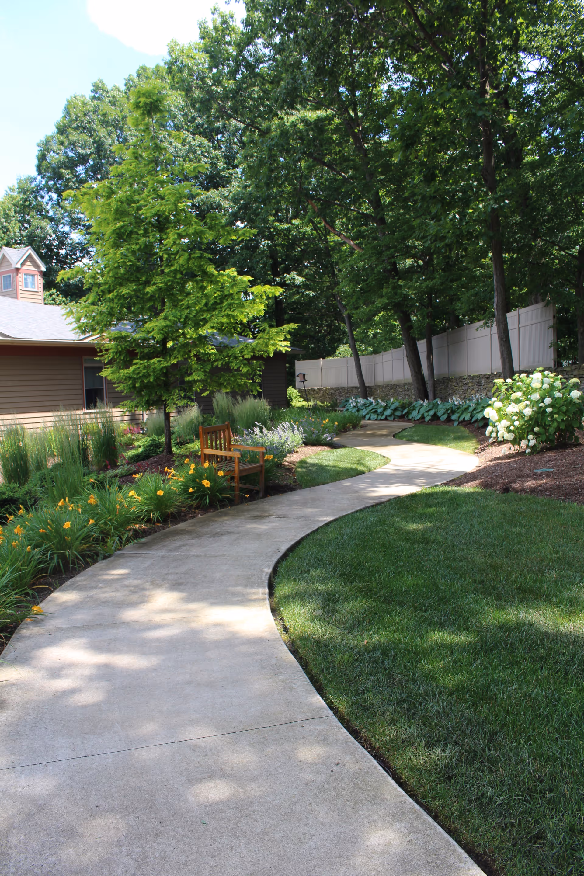 Curving concrete path through a landscaped garden with a wooden bench, lawn, flowers, and trees.