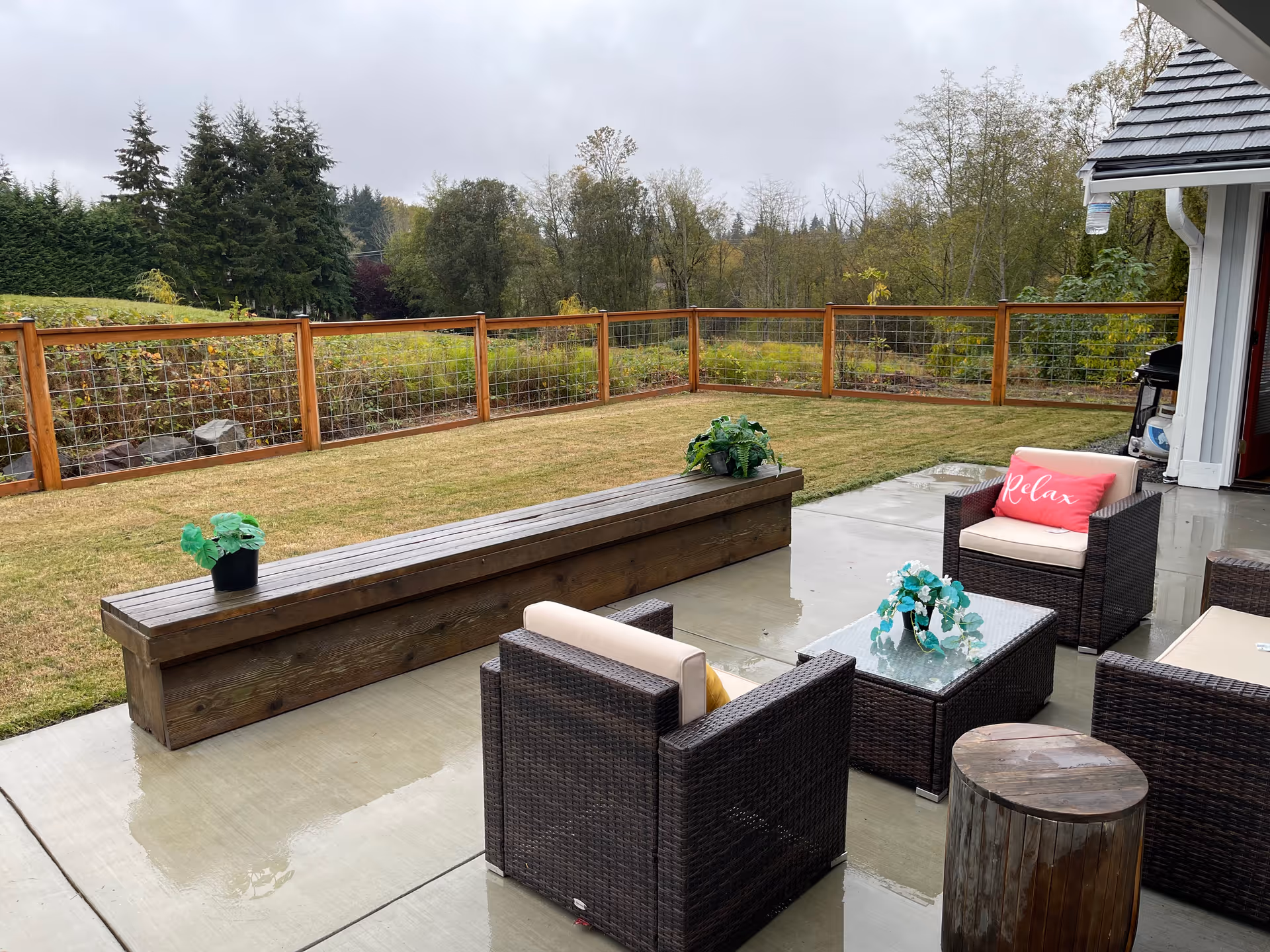 Outdoor patio area with wicker furniture including chairs and a glass-top coffee table, a wooden bench with potted plants, and a grassy yard enclosed by a wooden fence with wire mesh. Trees and greenery are visible in the background under a cloudy sky.
