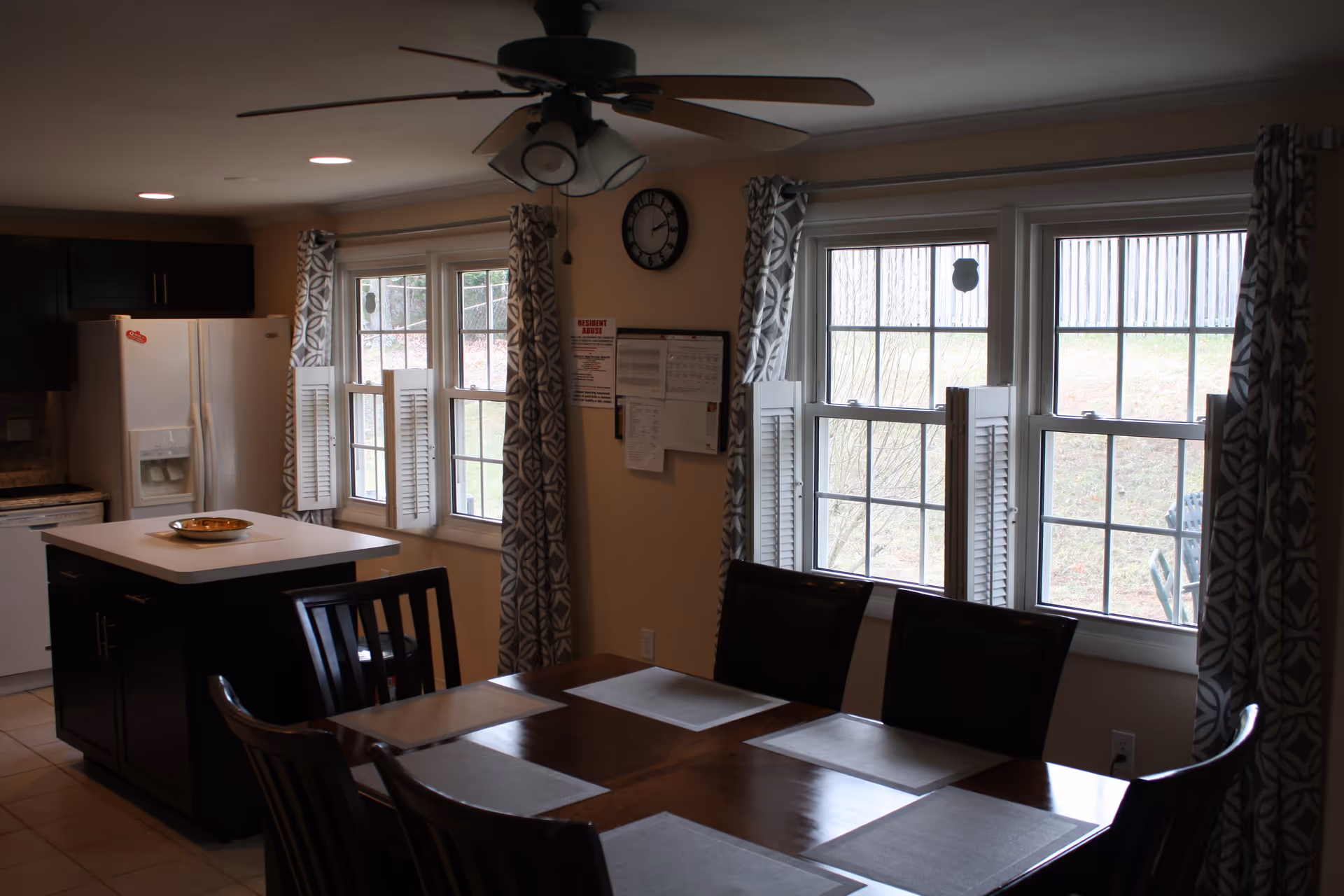 Interior view of a dining area and kitchen in an assisted living facility. The dining area features a wooden table with six chairs and placemats. The kitchen has a white refrigerator, dark cabinets, a kitchen island with a bowl on top, and windows with patterned curtains letting in natural light. A ceiling fan with lights is mounted above.