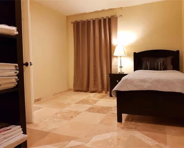 A bedroom with beige tiled floor, beige walls, and a dark wooden bed with white bedding and pillows. There is a nightstand with a lamp next to the bed, and brown curtains cover a window or door. On the left side, there is a shelving unit with neatly folded towels.