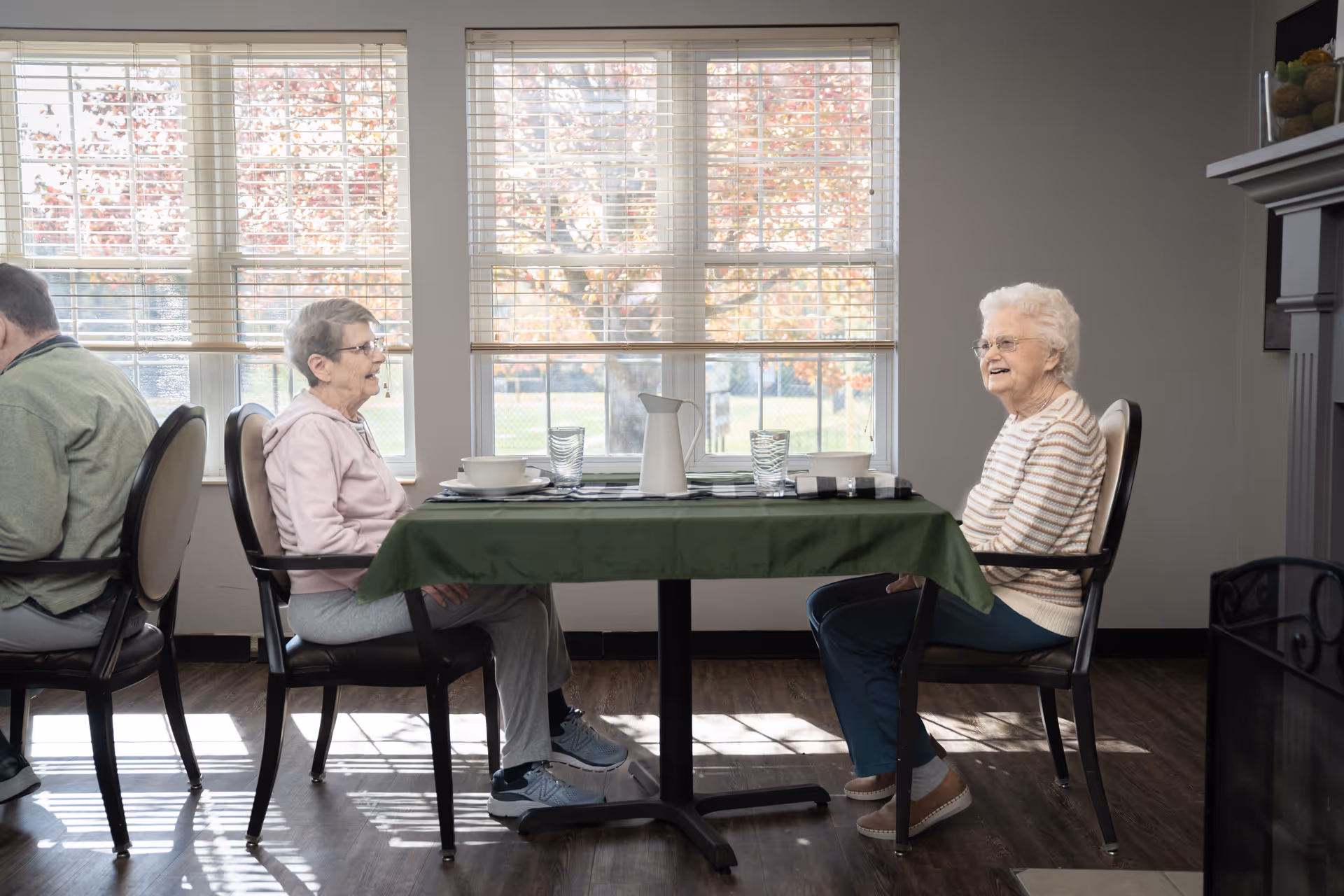 Two elderly women sitting at a dining table covered with a green tablecloth in a well-lit room with large windows showing autumn trees outside. The table is set with glasses, bowls, and a white pitcher. Another person is partially visible sitting on the left side of the image.
