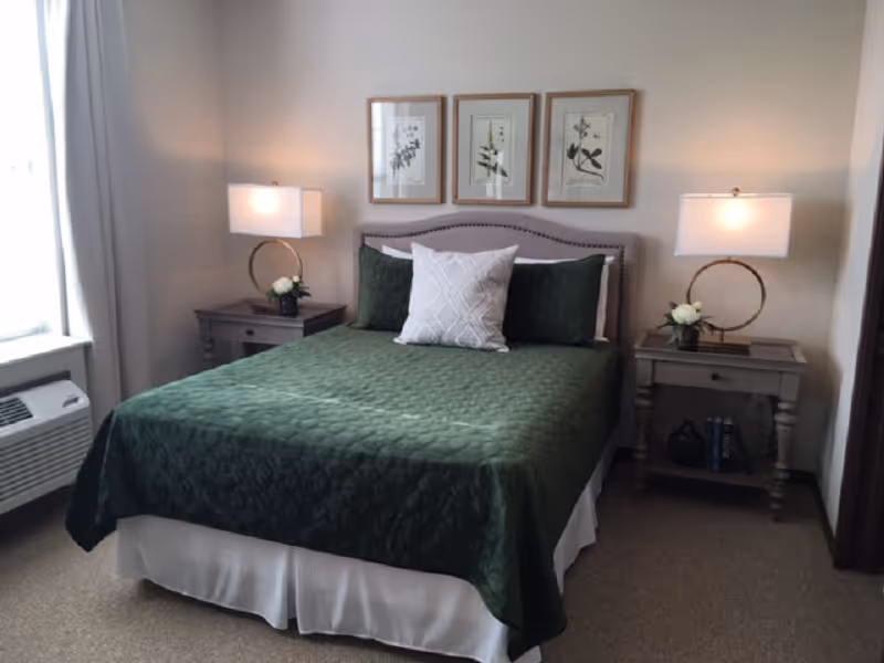 A neatly made bed with a green quilt, two bedside tables with lamps, and framed botanical prints on the wall in a bright bedroom.
