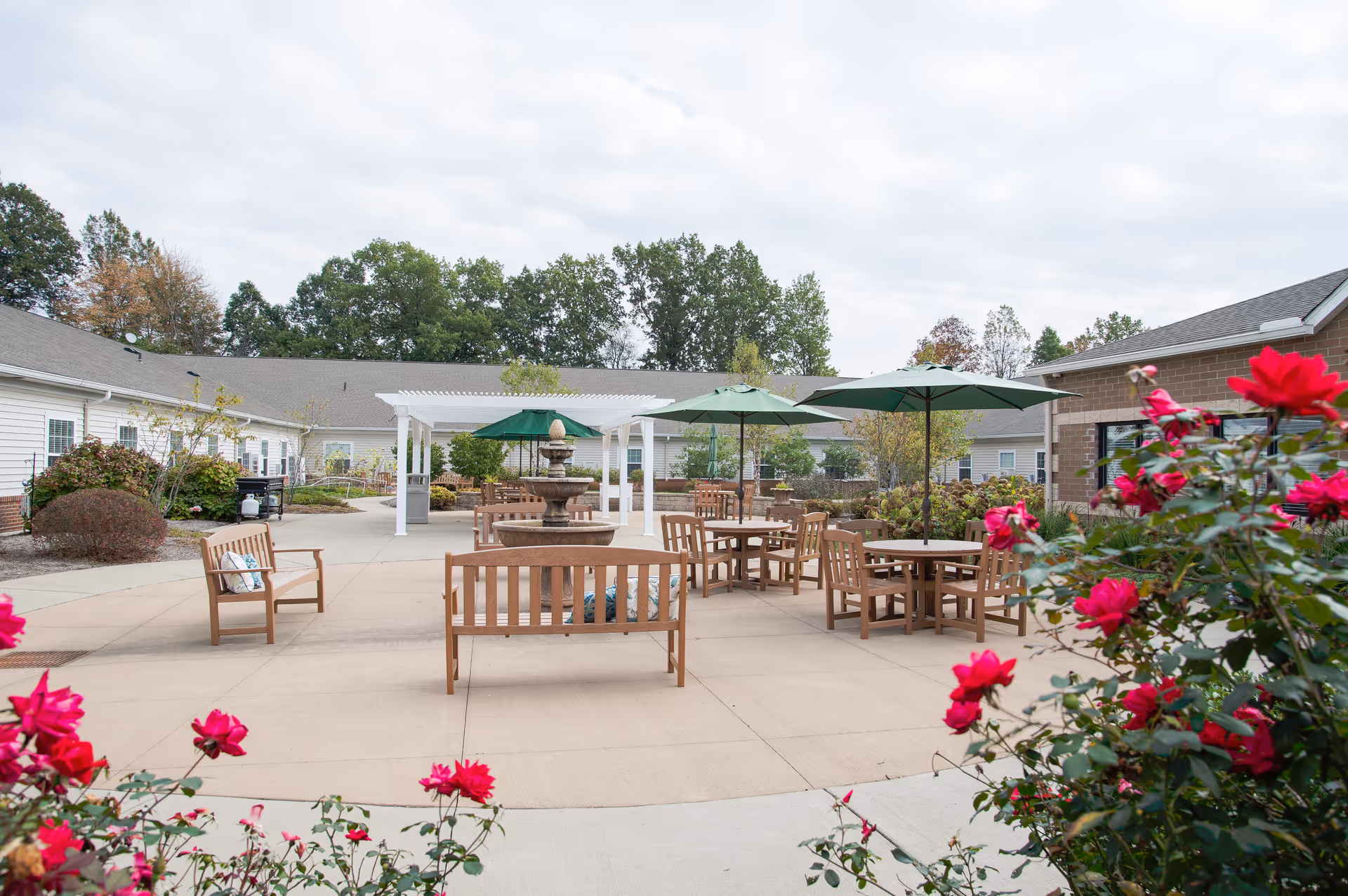 Outdoor courtyard area at Danbury Massillon featuring wooden benches and tables with green umbrellas, a central water fountain, surrounded by buildings and greenery with red flowers in the foreground under a cloudy sky.