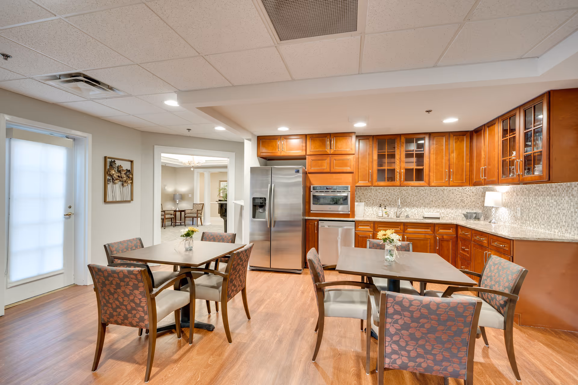 A bright kitchen and dining area in a senior living facility featuring wooden cabinets, stainless steel refrigerator and dishwasher, two square dining tables each surrounded by four cushioned chairs with patterned upholstery, and a door with frosted glass on the left side.