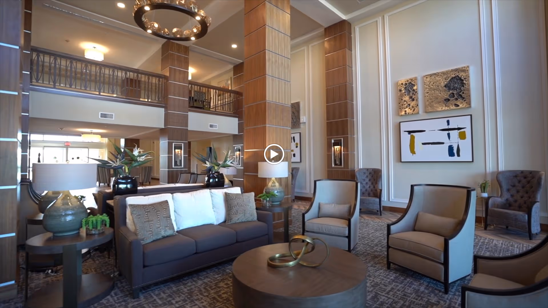 A spacious and elegant living room area with high ceilings and large wooden columns. The room features a dark gray sofa with white and patterned pillows, several beige armchairs, round wooden coffee tables with decorative items, and modern wall art. The space is well-lit with ceiling lights and lamps on side tables, and there is a balcony railing visible on the upper level.