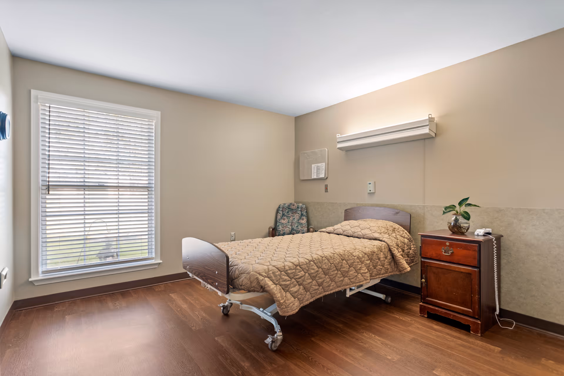 A simple, clean bedroom in a senior living facility with a single hospital-style bed covered with a beige quilted blanket. Next to the bed is a wooden nightstand with a small plant and a telephone. There is a floral upholstered chair in the corner and a large window with white blinds letting in natural light. The walls are painted beige with a darker beige wainscoting, and the floor is wood.