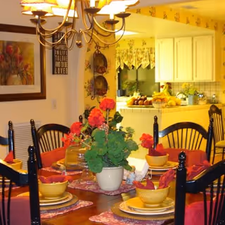 A warmly lit dining room with a wooden table set for six people. The table features yellow bowls, plates, and red napkins, with a centerpiece of red flowers in a white pot. In the background, a kitchen area with white cabinets and a countertop filled with fruit is visible. The walls are decorated with floral wallpaper and framed artwork.